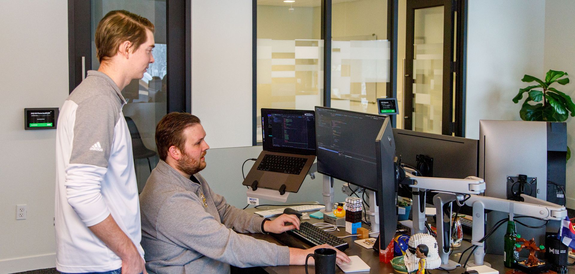 Two male developers review work at a desk with multiple computer screens